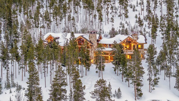 A large, multi-story lodge with a stone facade and wooden exterior, surrounded by snow-covered ground and tall pine trees.
