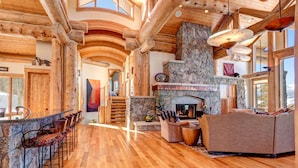 A spacious log cabin interior with a stone fireplace, wooden beams, and a large window overlooking a snowy landscape.