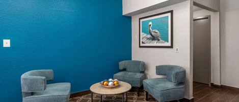 A room with a blue wall, a framed painting of a pelican, and three grey armchairs around a round table with a bowl of fruit.