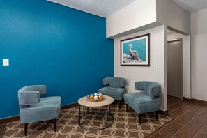 A room with a blue wall, a framed painting of a pelican, and three grey armchairs around a round table with a bowl of fruit.