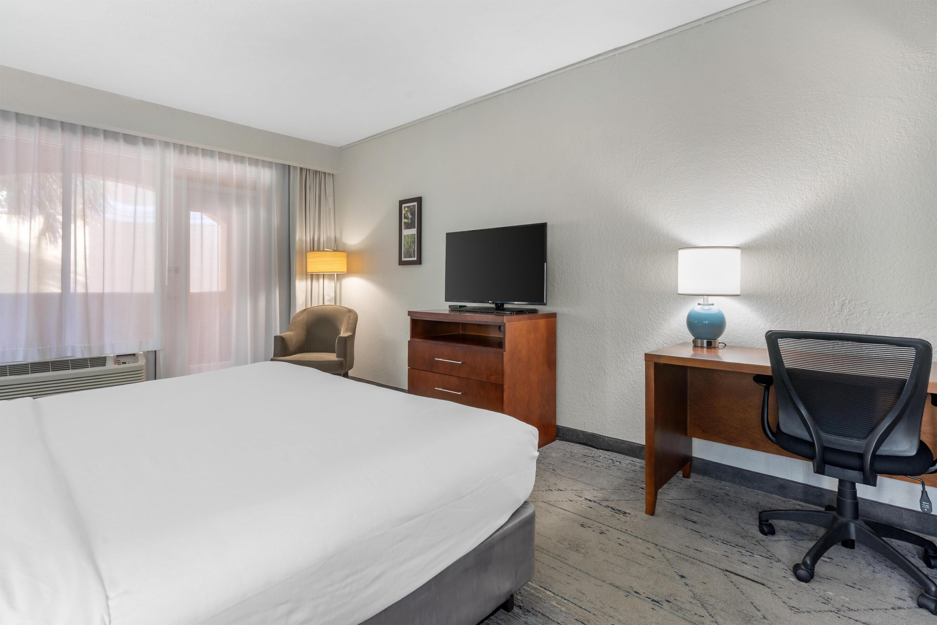 A hotel room with a bed, a desk with a chair, a television, and a lamp.