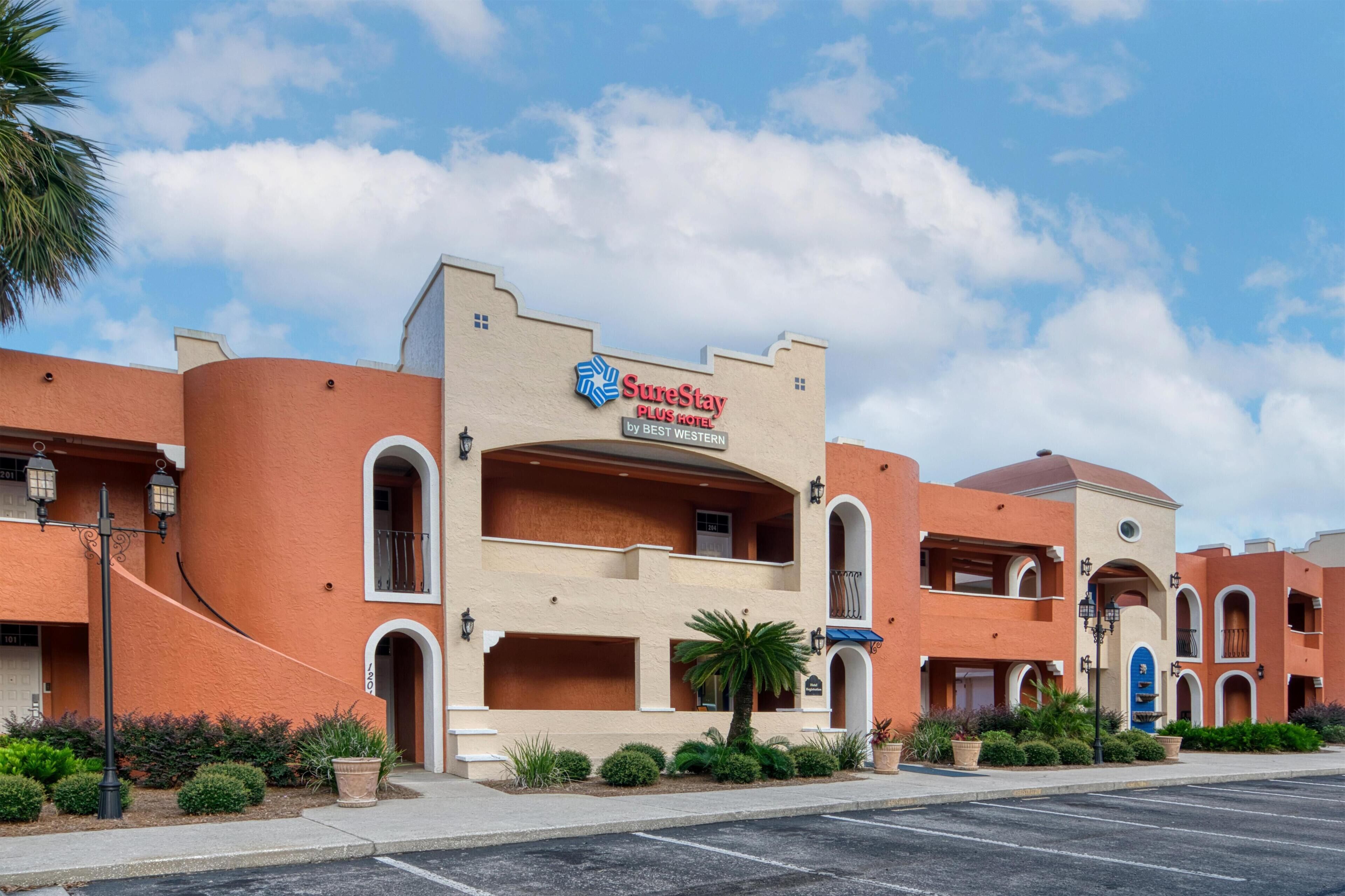 A hotel with a 'SureStay Plus Hotel by Best Western' sign, featuring a stucco exterior, arched windows, and a palm tree in the foreground.