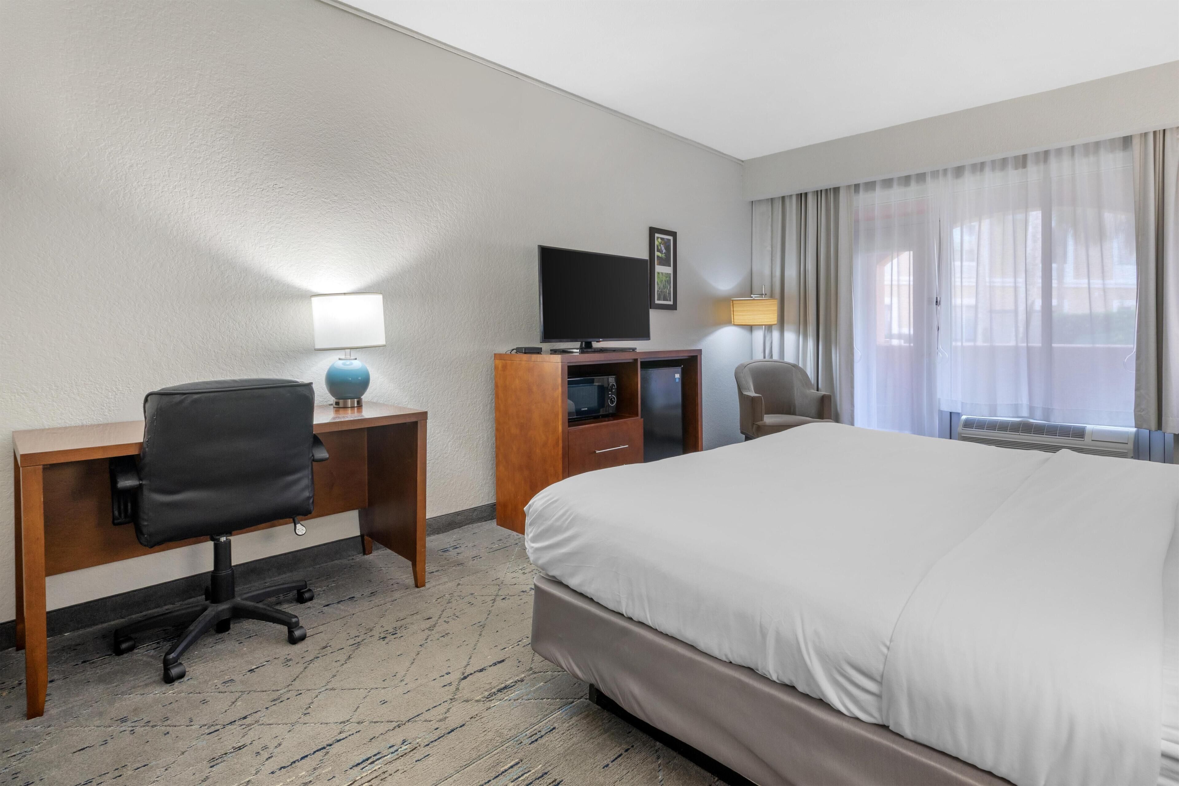 Habitación de hotel con cama, escritorio con silla, televisor y ventana con cortinas.