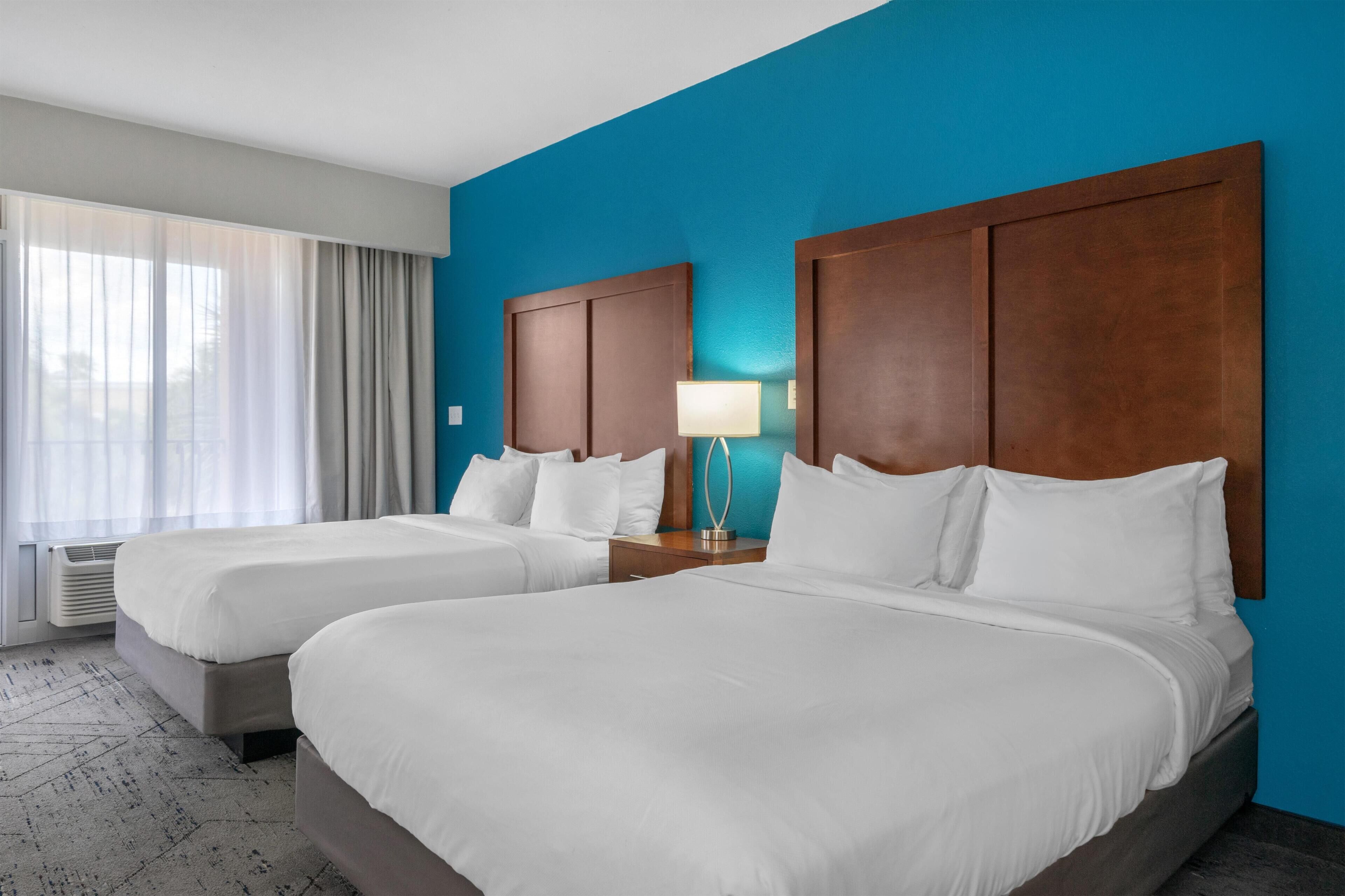 A hotel room with two beds, a nightstand with a lamp, and a view of the outdoors through a window with curtains.