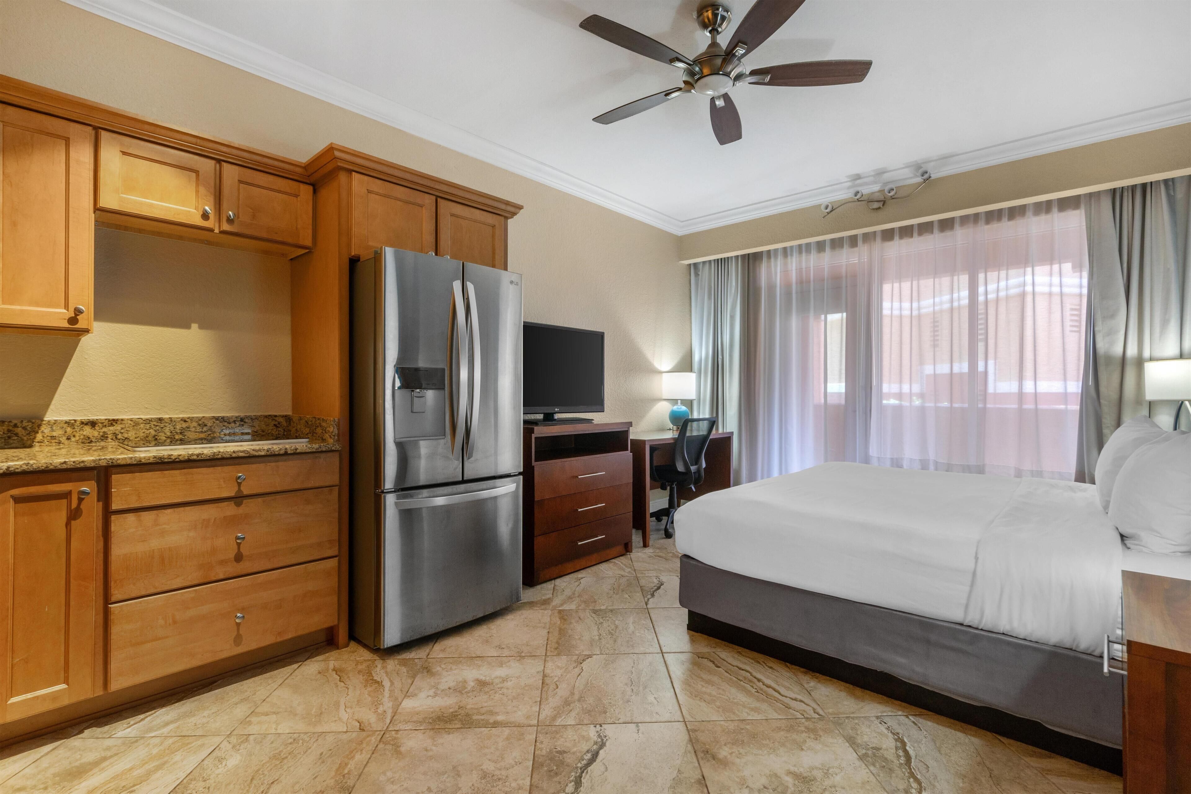 A hotel room with a bed, a desk with a chair, a television, a refrigerator, wooden cabinets, and a ceiling fan.