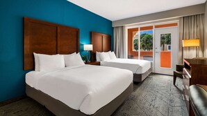 Une chambre d’hôtel avec deux lits, une tête de lit en bois, une table de chevet avec une lampe, un bureau avec une chaise et une vue sur des palmiers à travers une porte vitrée.