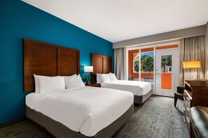 A hotel room with two beds, a wooden headboard, a nightstand with a lamp, a desk with a chair, and a view of palm trees through a glass door.