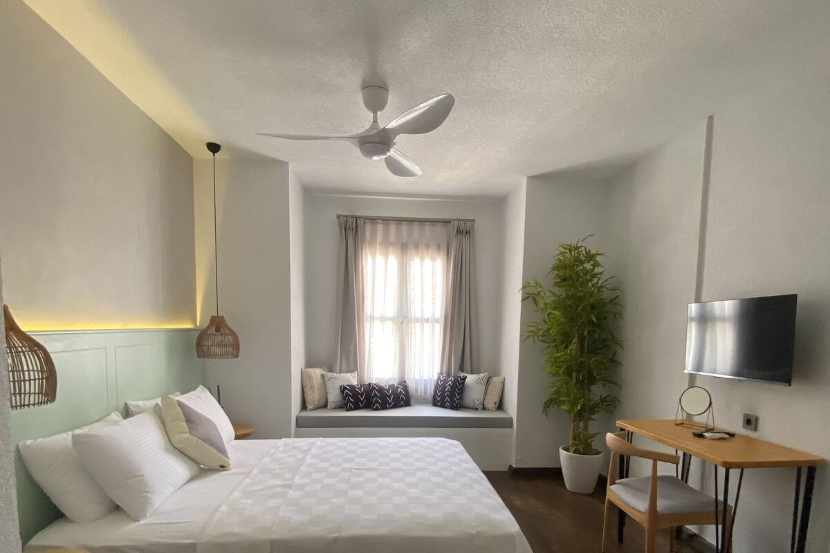 A bedroom with a bed, a desk, a chair, a ceiling fan, a window with curtains, and a potted plant.
