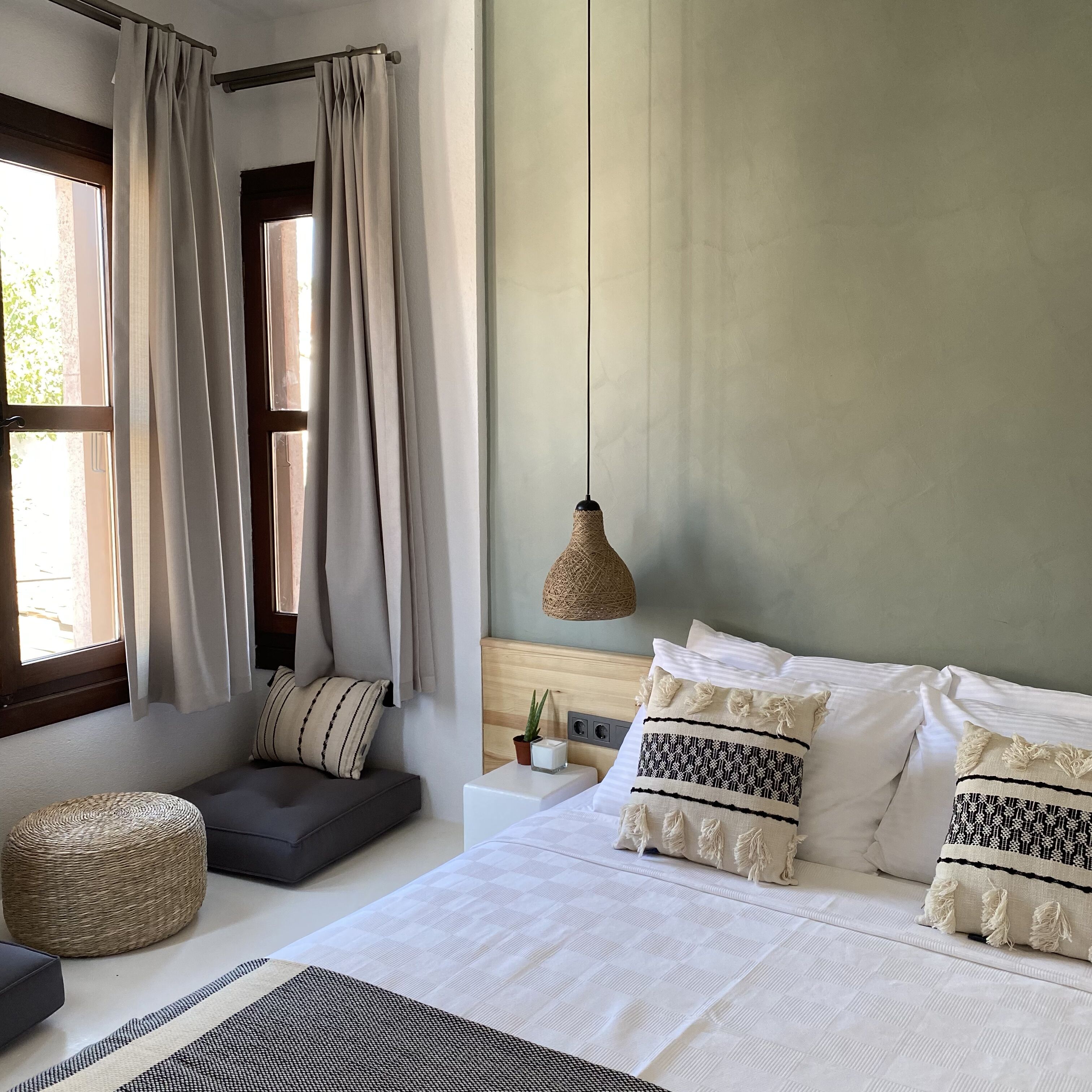 A bedroom with a bed, bedside table, curtains, a hanging light fixture, and a woven ottoman.