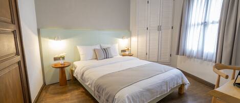 Un dormitorio con cama, mesitas de noche, una silla, armario y ventana con cortinas.