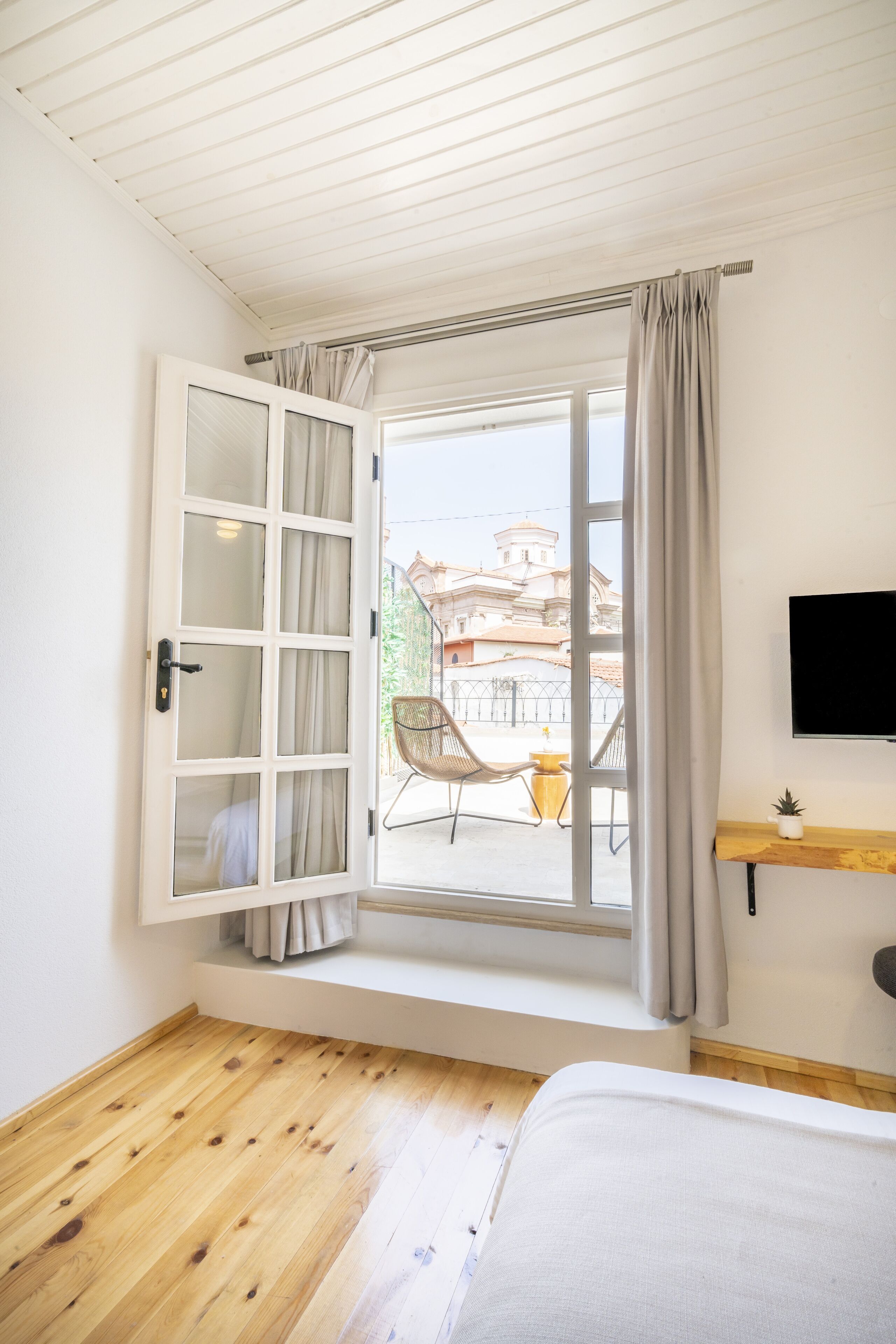 Una habitación con piso de madera, una cama, una ventana con cortinas, una silla y un televisor en la pared.