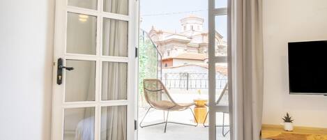 Una habitación con piso de madera, una cama, una ventana con cortinas, una silla y un televisor en la pared.