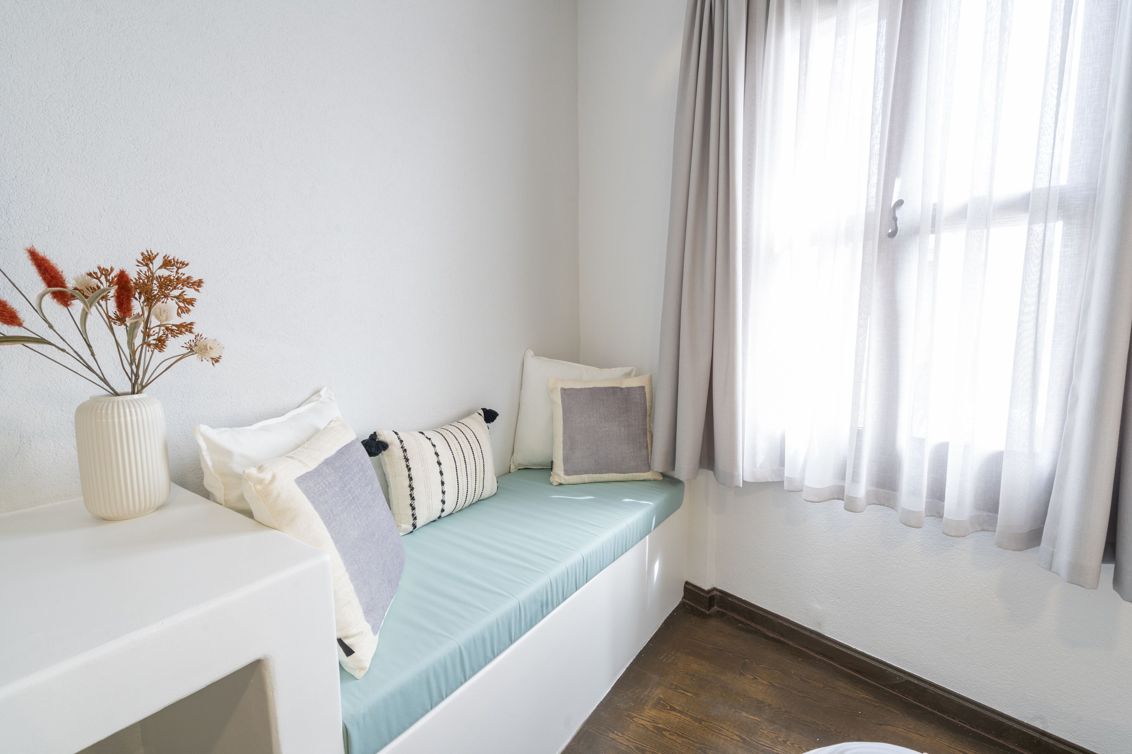 Una habitación con cama, una mesita de noche, un florero con flores secas y una ventana con cortinas.