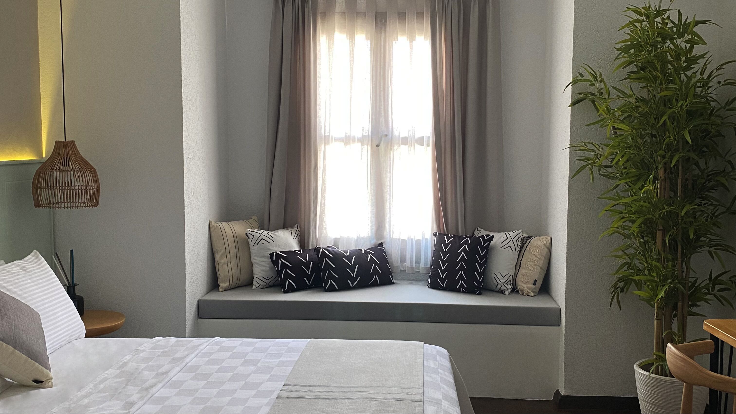 A bedroom with a bed, a window seat with cushions, a hanging lamp, and a potted plant.