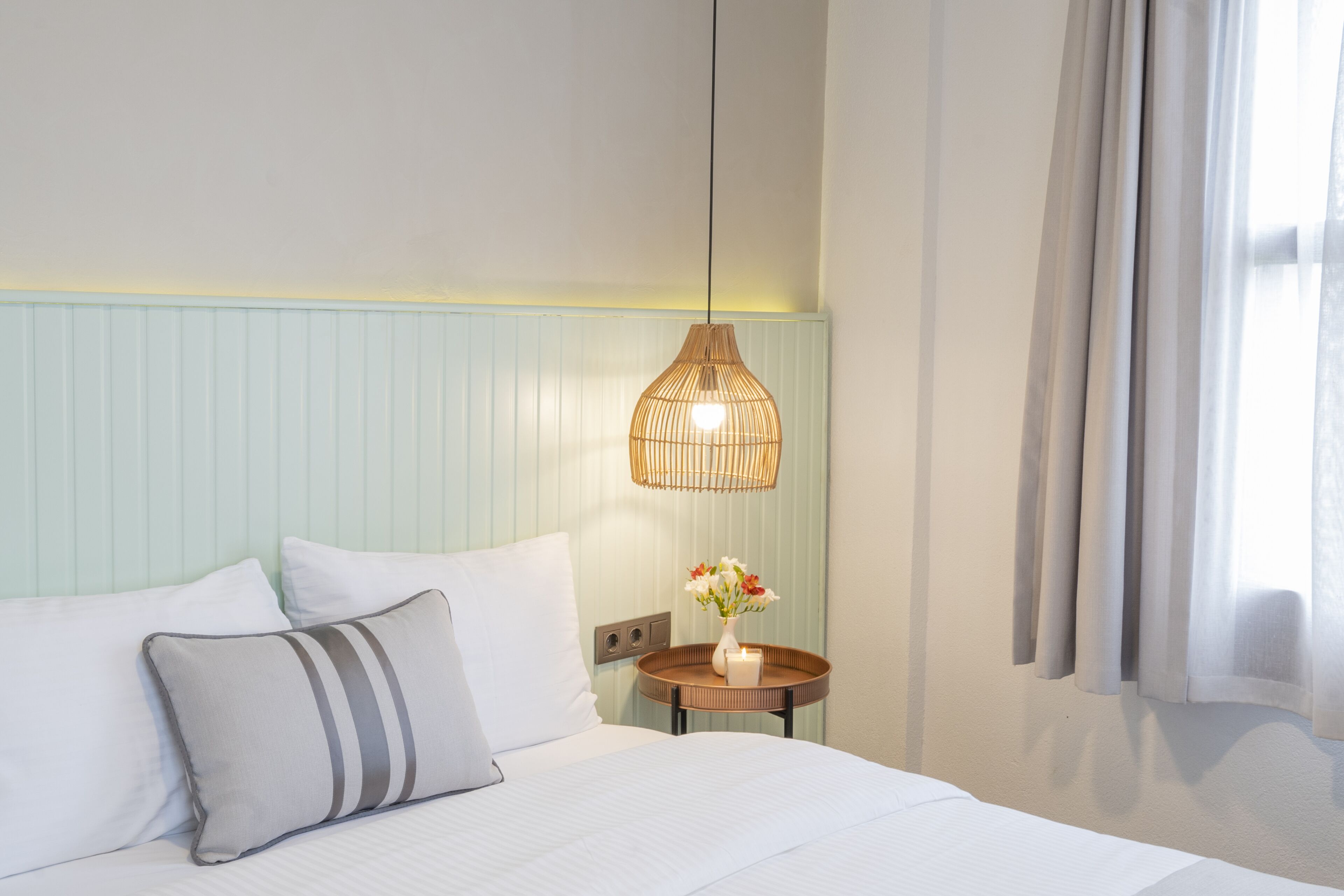 Un lit bien fait, avec du linge de lit blanc et des oreillers à rayures, une table de chevet ornée d’un vase de fleurs et un luminaire suspendu.