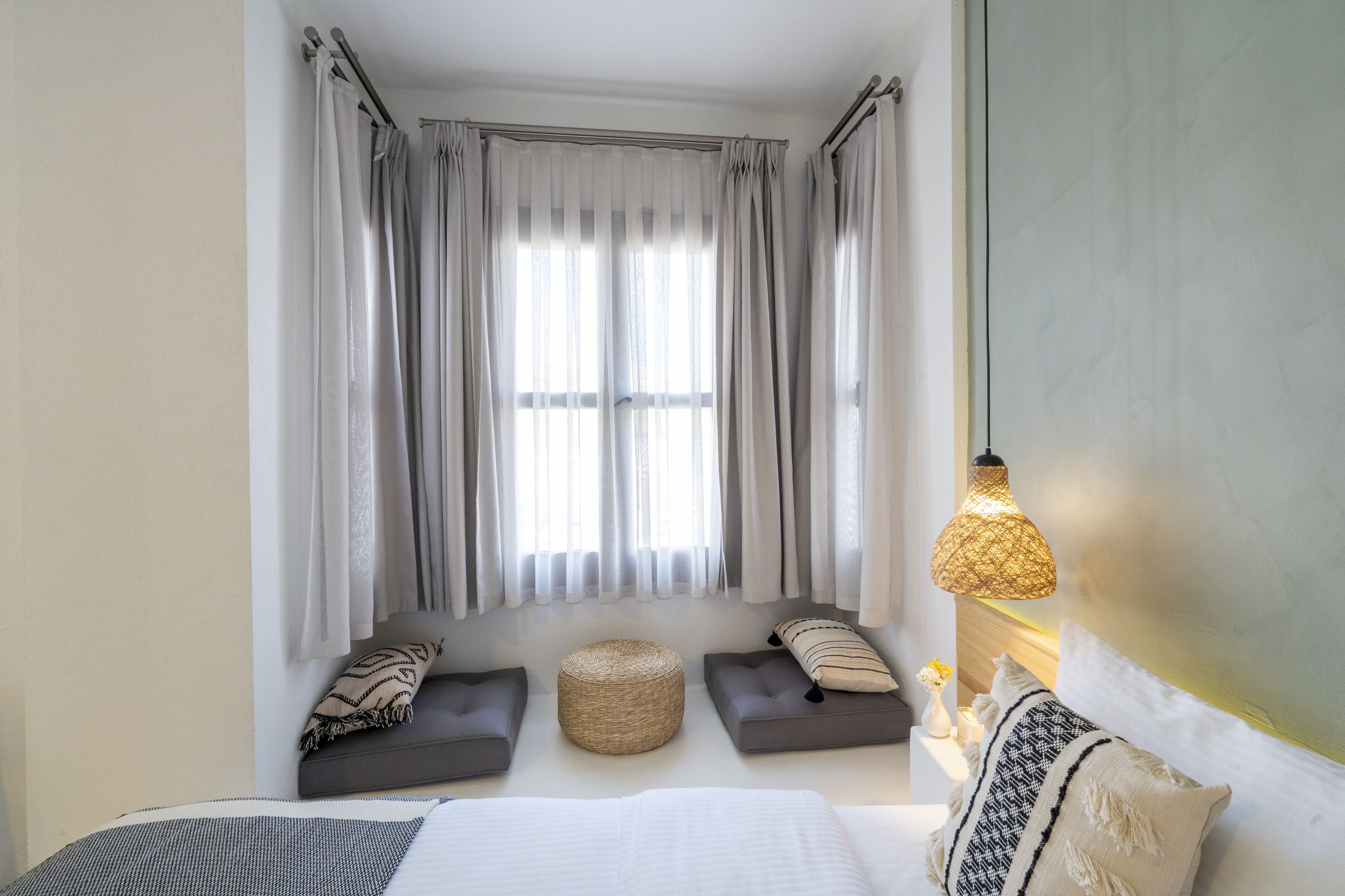 Quarto com cama, dois sofás cinzas, um banquinho trançado e uma mesinha de cabeceira com abajur.