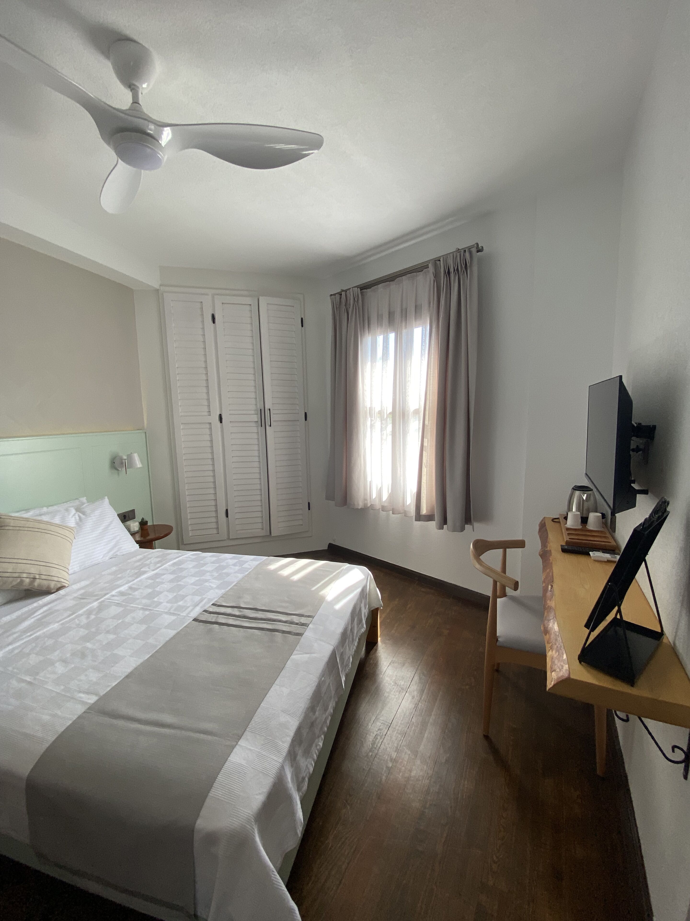 A hotel room with a bed, a desk with a computer, a ceiling fan, and a window with curtains.