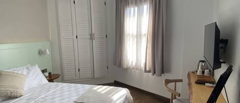 A hotel room with a bed, a desk with a computer, a ceiling fan, and a window with curtains.