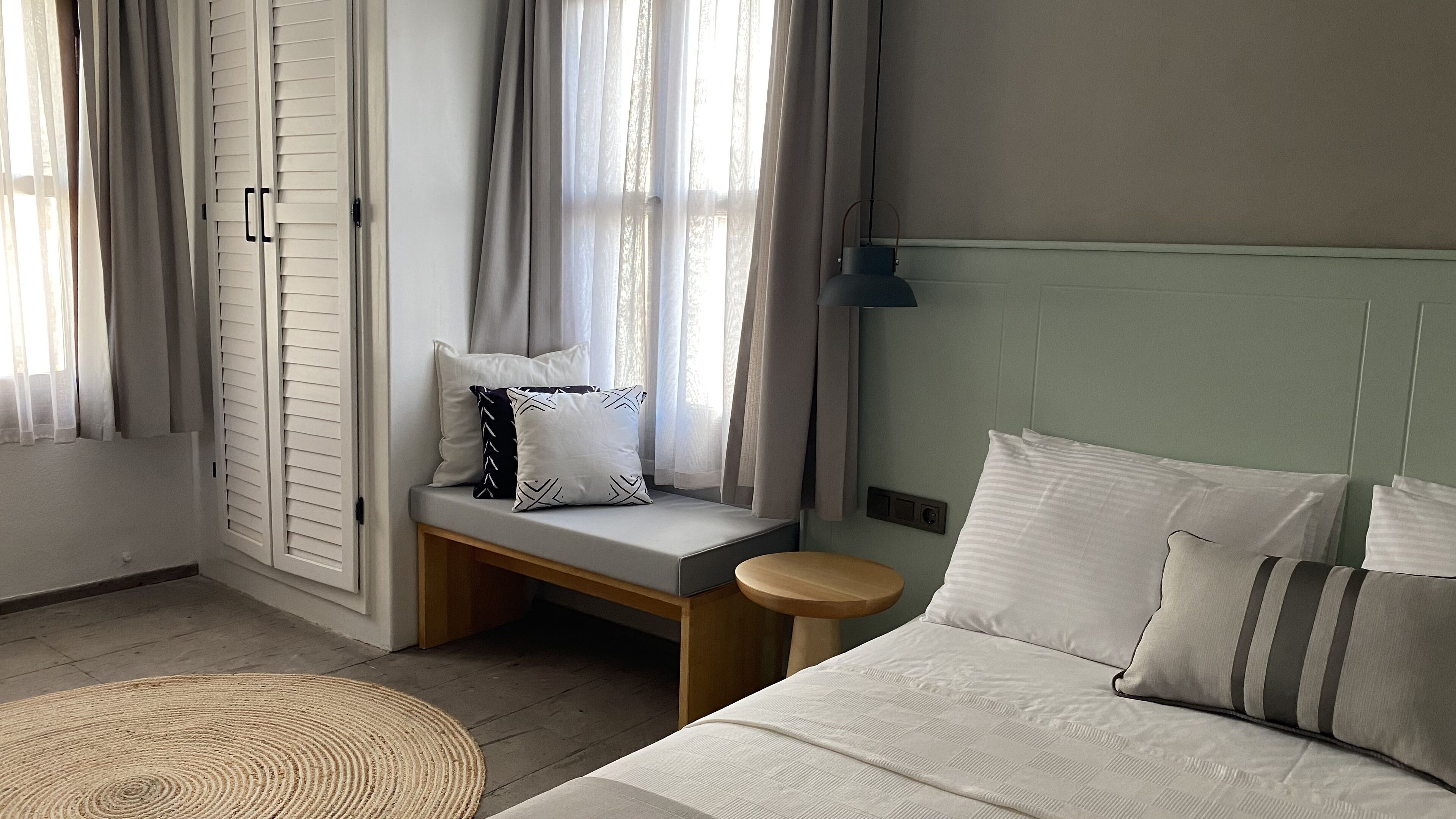 A bedroom with a bed, a bench with pillows, a bedside table, and a window with curtains.