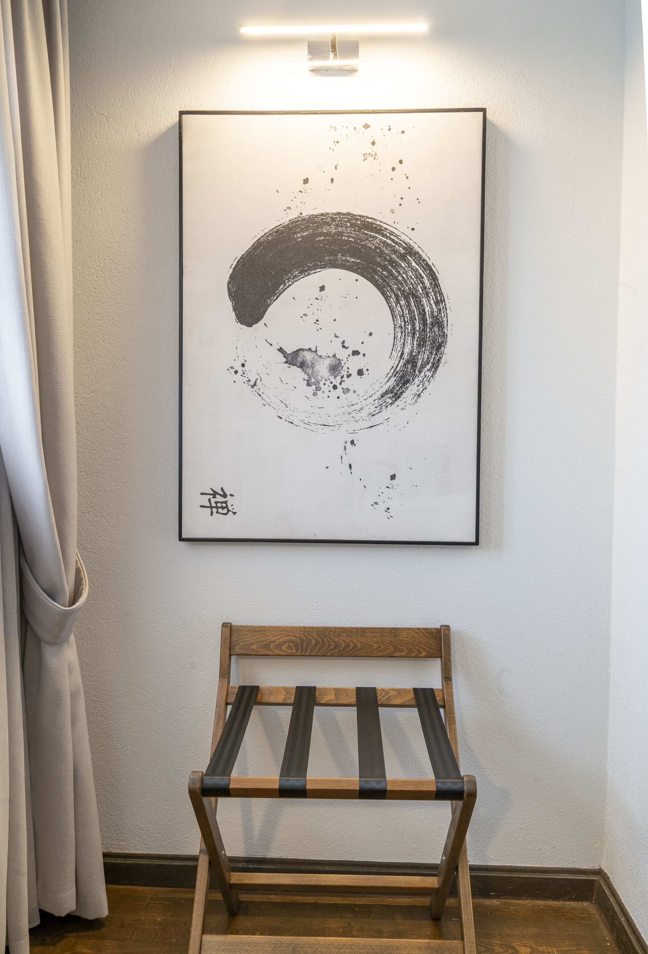 A framed abstract artwork on a wall, a wooden chair in front, and a curtain partially covering a window.