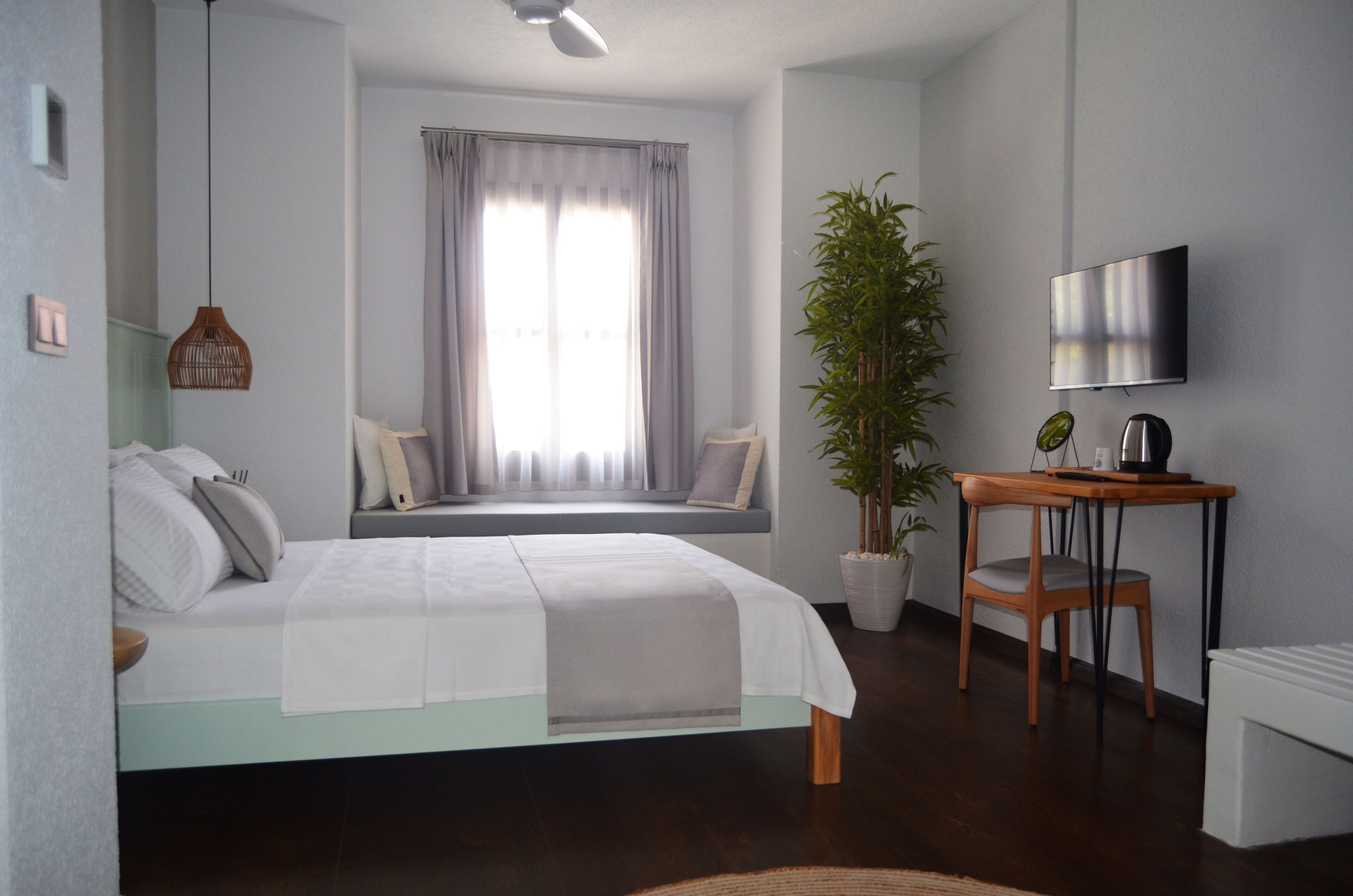A modern bedroom with a bed, a window with curtains, a desk with a chair, a potted plant, and a flat-screen TV on the wall.