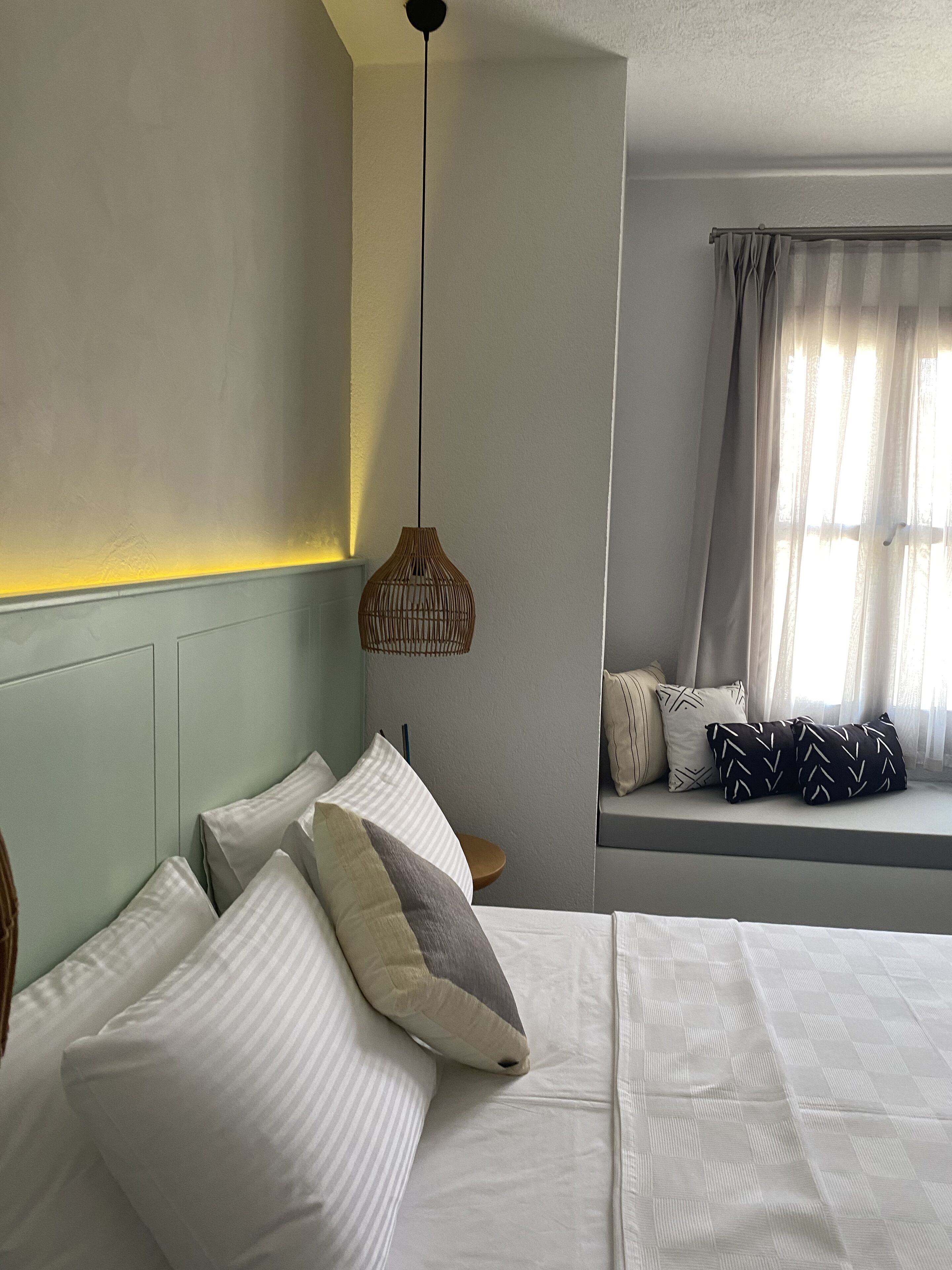 A bedroom with a bed, pillows, a bedside table, a hanging light fixture, and a window with curtains.
