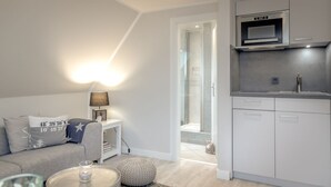 A modern living room with a grey sofa, a small round table, and a lamp. There is a kitchen area with white cabinets and a microwave. A glass shower enclosure is visible in the background. - Ben, Tinnum, Germany (Sylt-Ost)