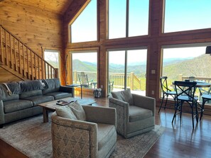 A spacious living room with a wooden interior, a sofa, armchairs, a coffee table, and a view of the mountains through large windows.