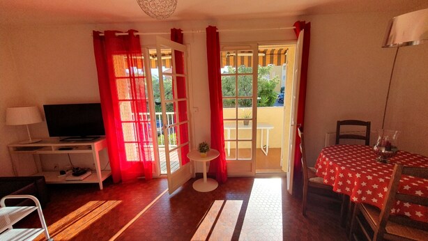 A room with a TV on a stand, a small table with a plant, a dining table with a star-patterned tablecloth, and a balcony with red curtains. - South-facing T2 apartment in village center with parking, swimming pool and tennis court. (La Croix Valmer)
