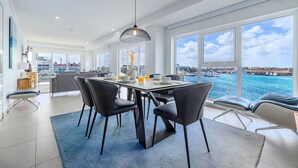 A modern dining area with a large table, chairs, and a view of the sea through large windows.