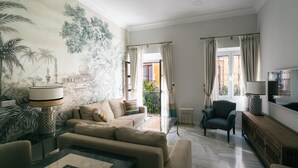 A living room with a sofa, armchairs, a coffee table, and a TV. There is a mural on the wall and a window with curtains. - Apartamentos Abreu Suites (Seville)