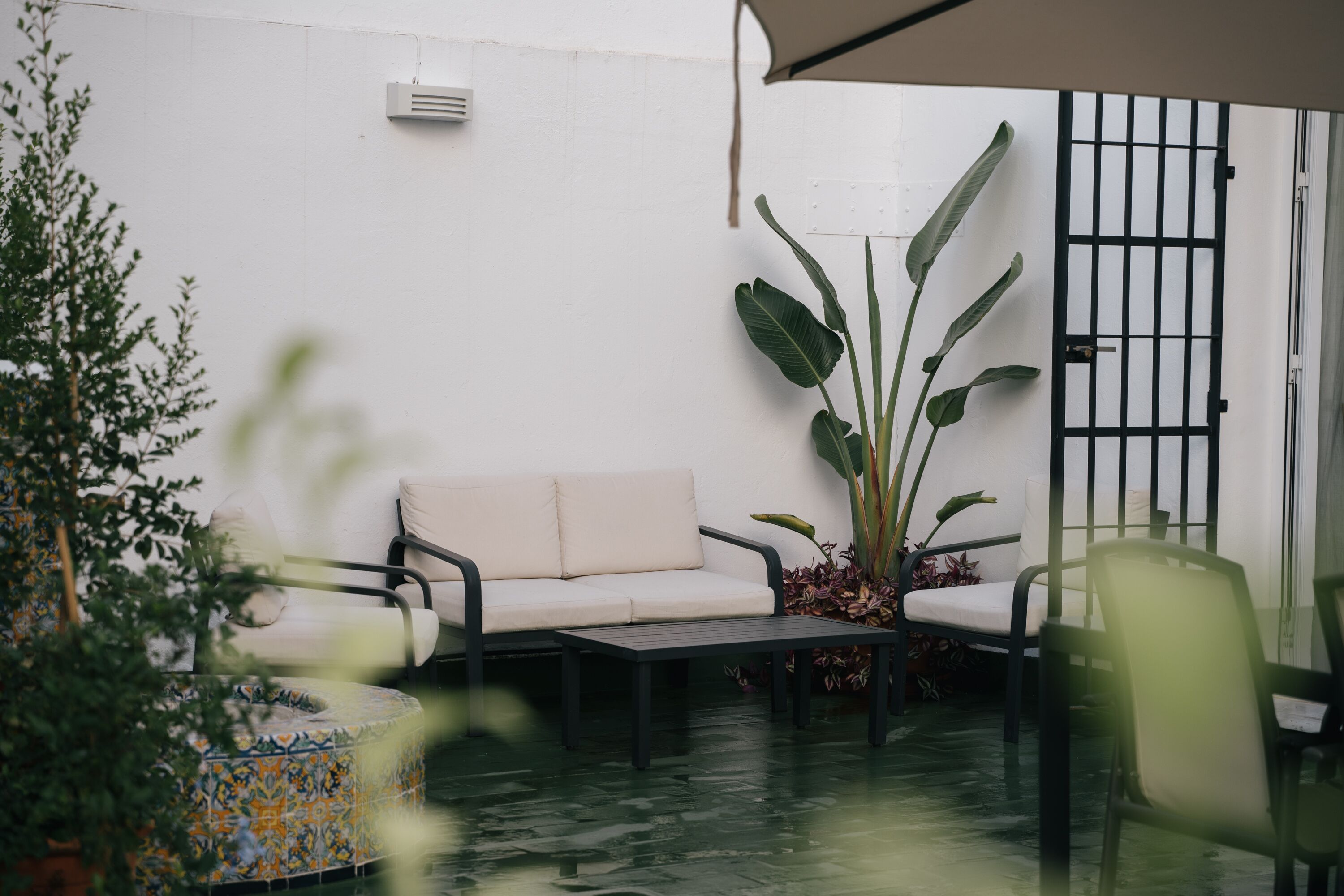 Un patio con un sofá blanco, una mesa redonda y sillas. Se observa una planta grande y un piso con baldosas.