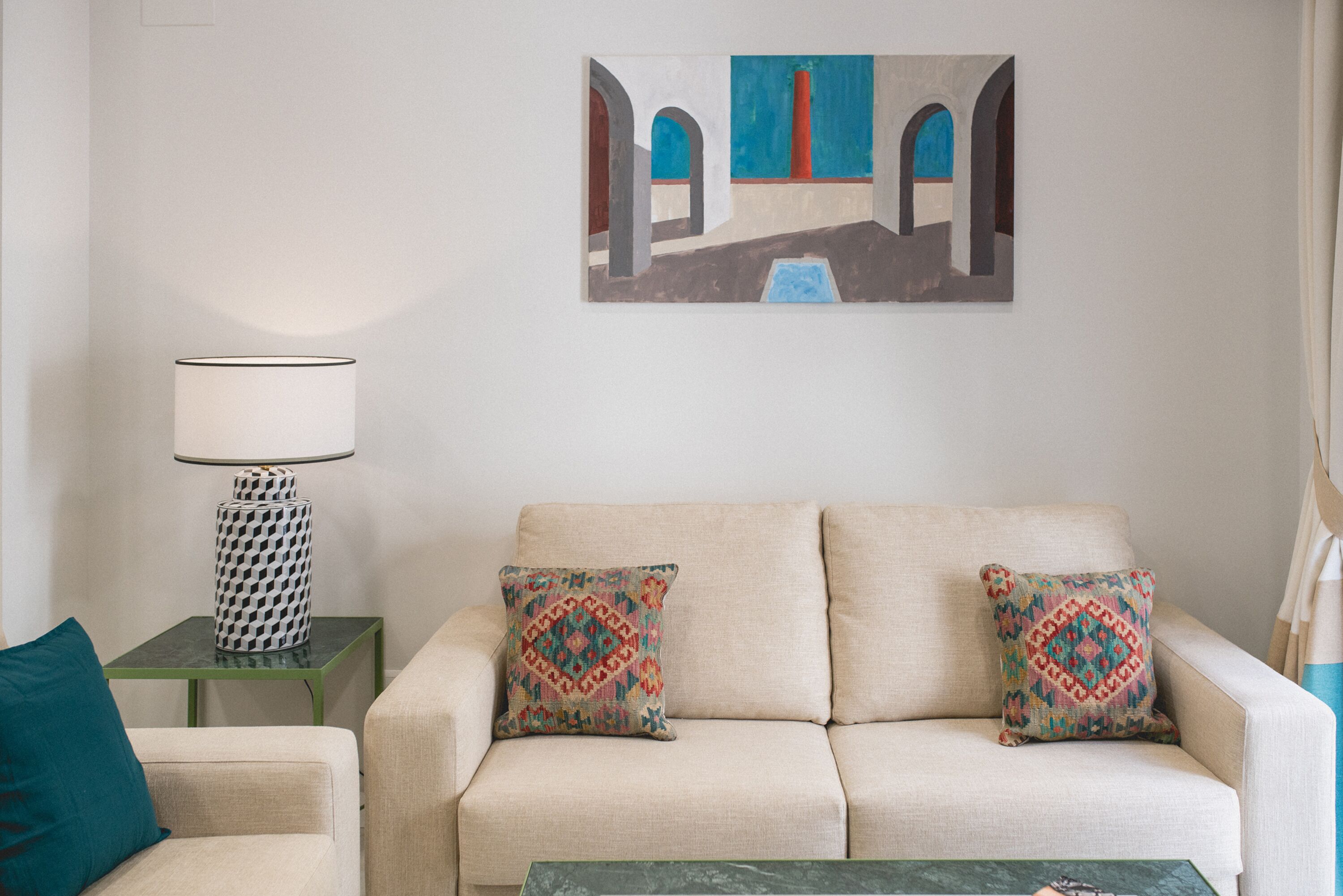 Una sala de estar con un sofá color beige, dos almohadones estampados, una mesita auxiliar verde con una lámpara, y un cuadro con una escena arquitectónica en la pared.