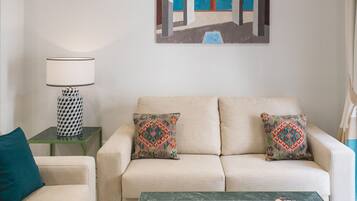 Una sala de estar con un sofá color beige, dos almohadones estampados, una mesita auxiliar verde con una lámpara, y un cuadro con una escena arquitectónica en la pared.