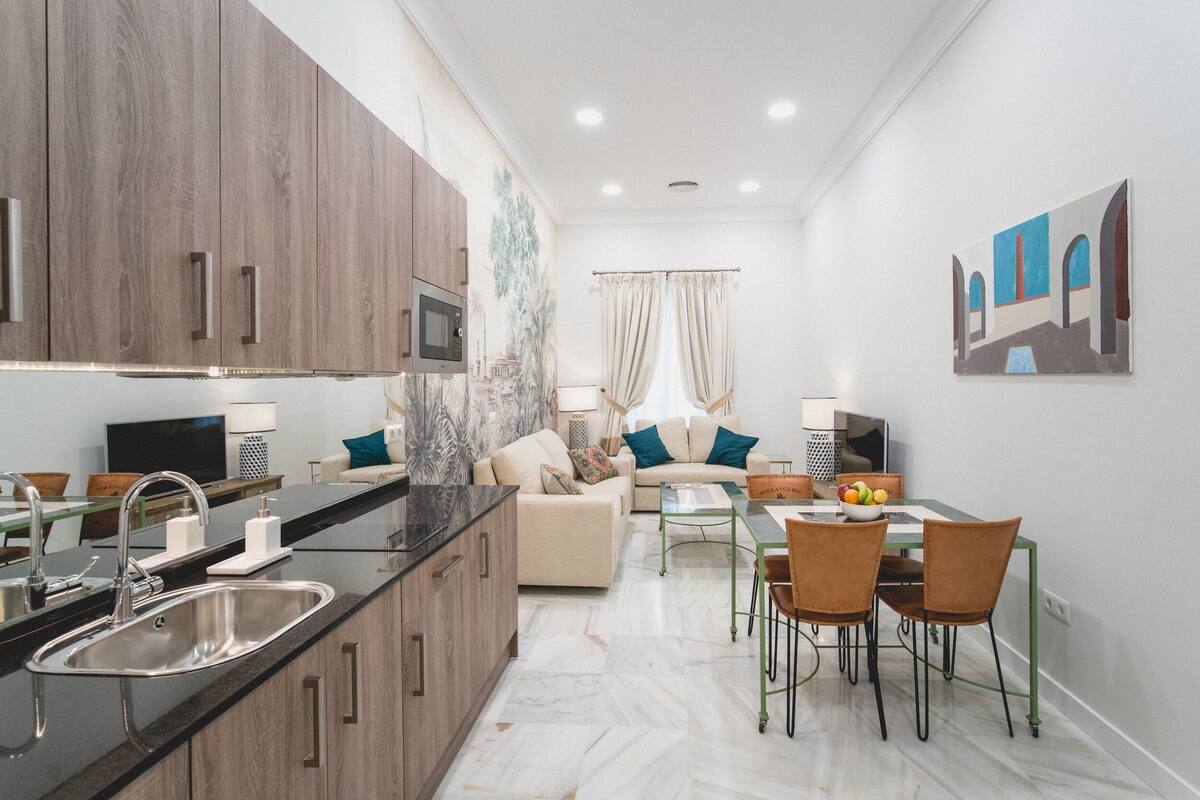 A modern kitchen with a marble countertop, stainless steel sink, and wooden cabinets. There is a dining area with a glass table and chairs, and a living area with a sofa and curtains.