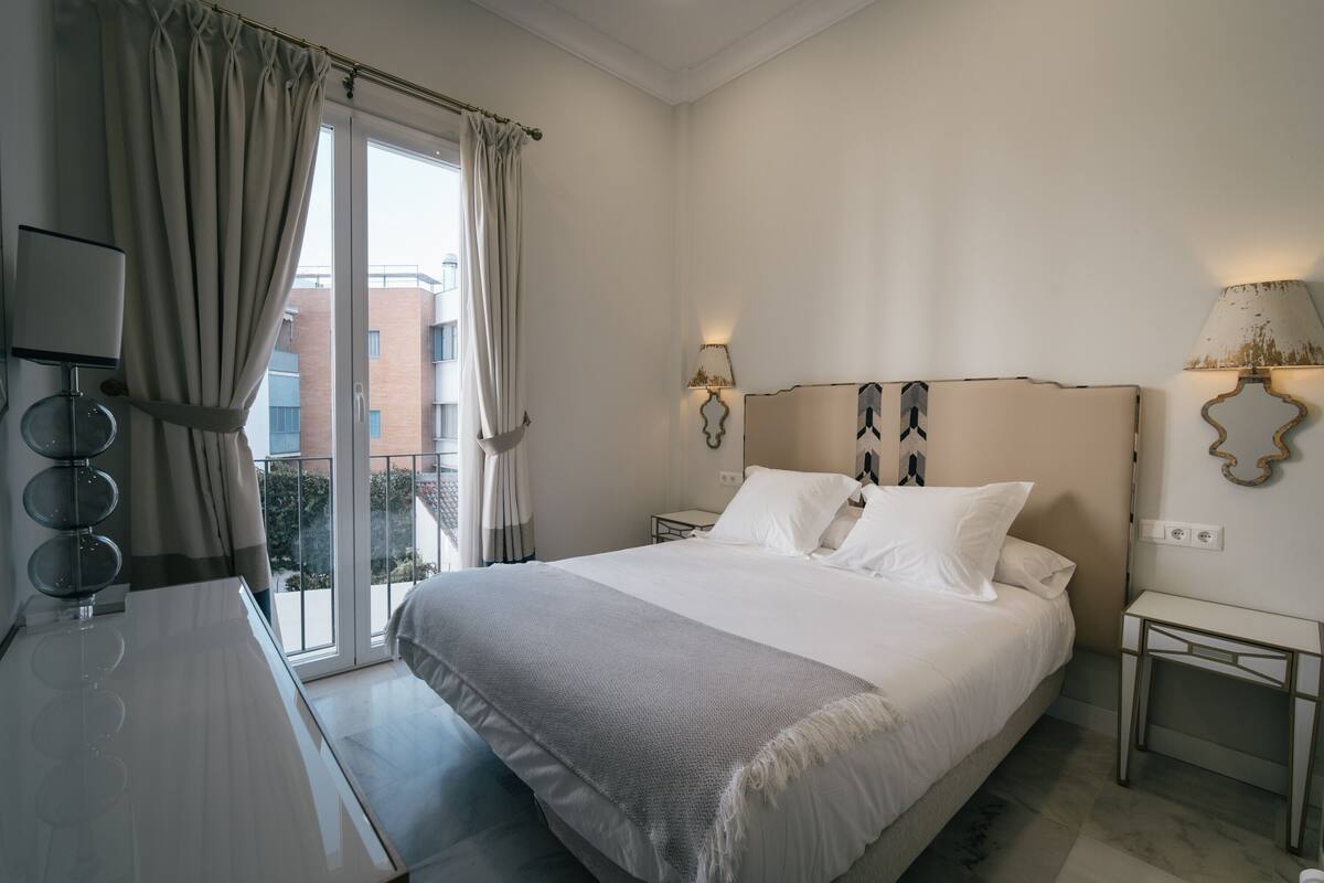 A hotel room with a bed, bedside lamps, a desk, and a view of buildings through the window.