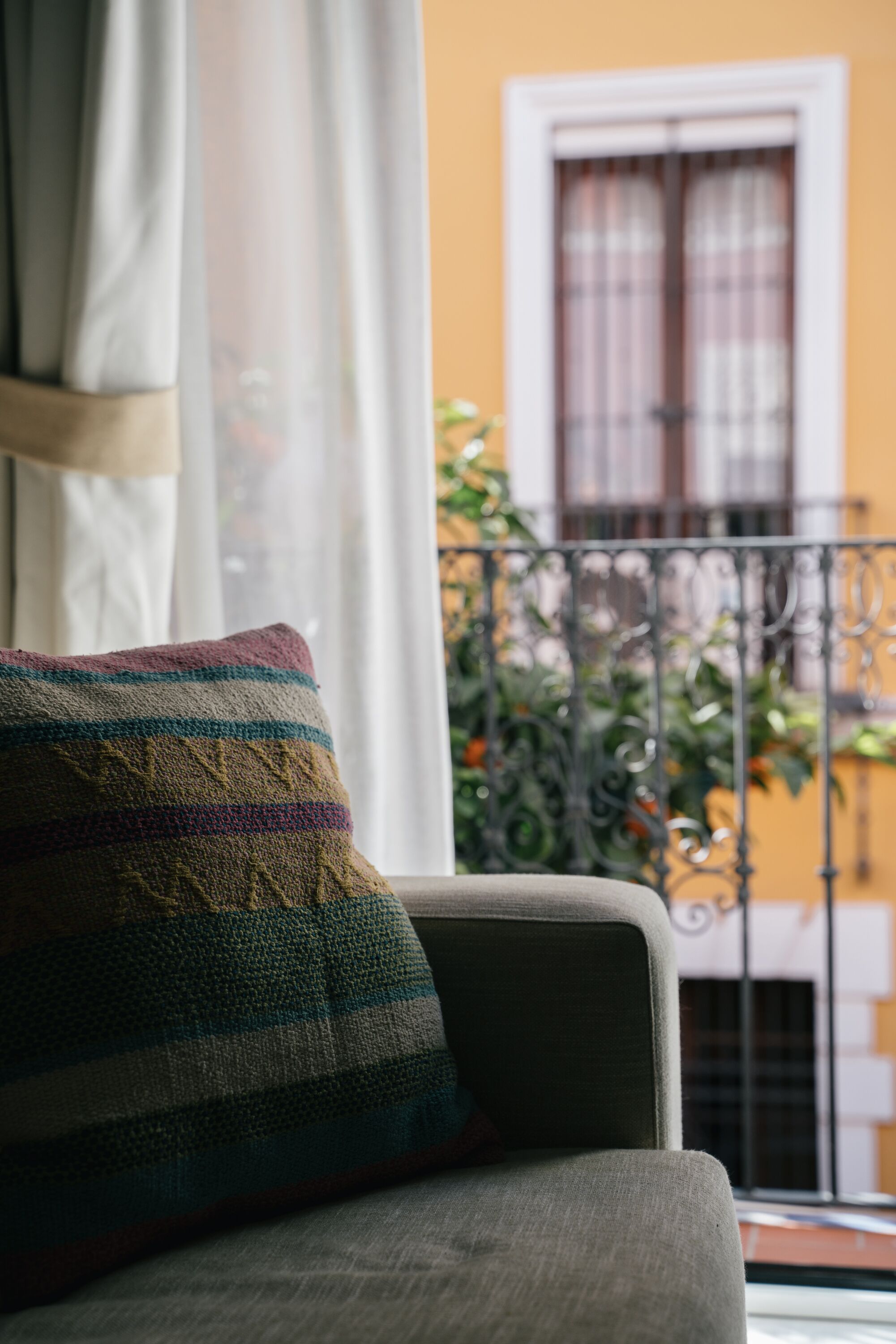 Una habitación con sofá, un cojín estampado, una ventana con cortinas y un balcón con barandilla de metal y plantas.