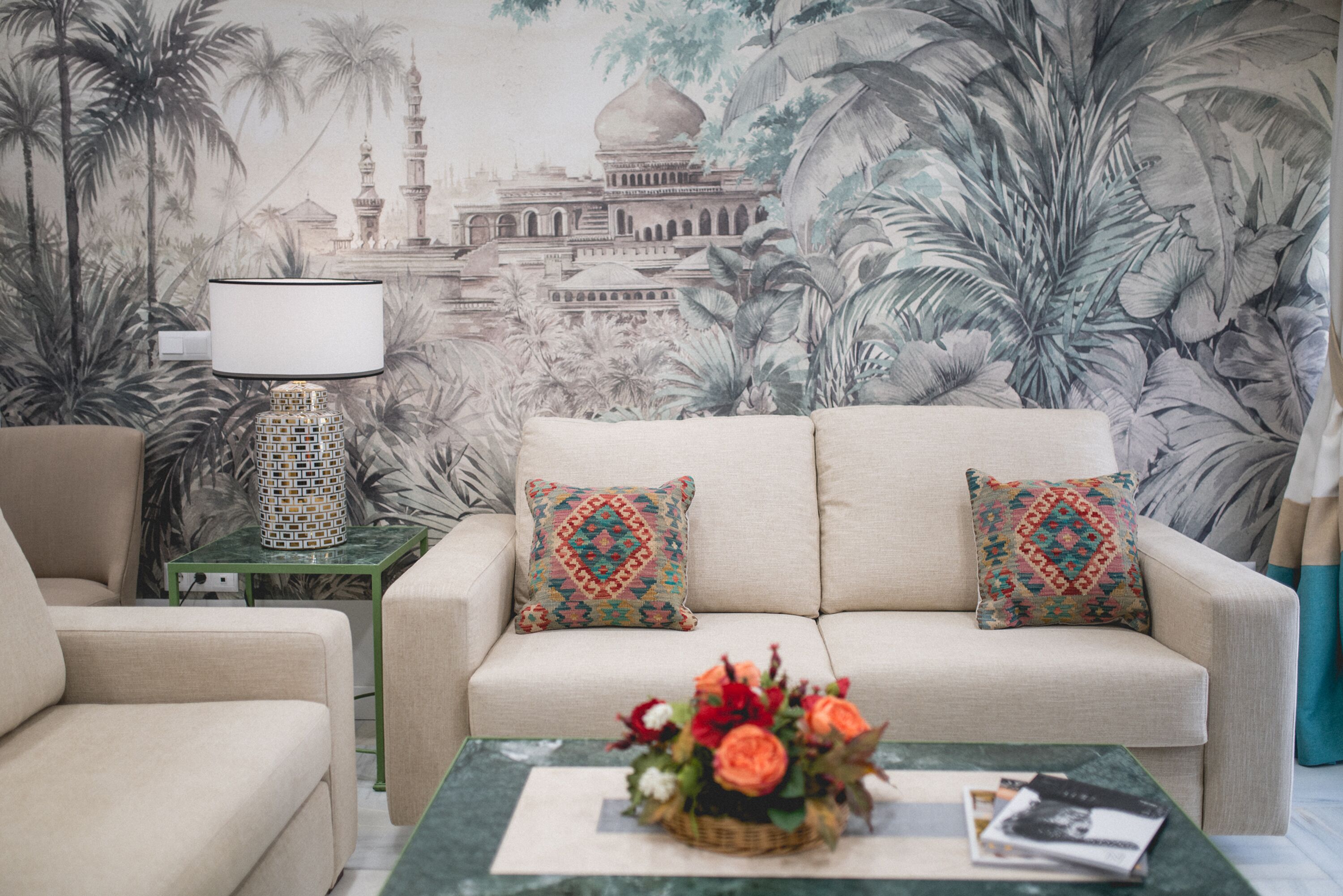 Ein Wohnzimmer mit einem beigen Sofa, gemusterten Kissen, einem grünen Couchtisch mit floraler Dekoration und einer Wandmalerei, die ein Gebäude und tropische Pflanzen zeigt.