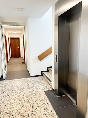 Ein Flur mit einer Holztür am Ende, einer Holztreppe mit Geländer und einem modernen Aufzug.