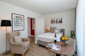 Ein Hotelzimmer mit Bett, Sessel, Tisch mit Zeitschrift und Wein, Lampe sowie Wand mit alphabetischem Aushang und Weinausstellung.