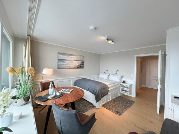 A modern bedroom with a bed, bedside tables, a desk, and a chair. There is a window with curtains, a painting on the wall, and a door leading to another room.
