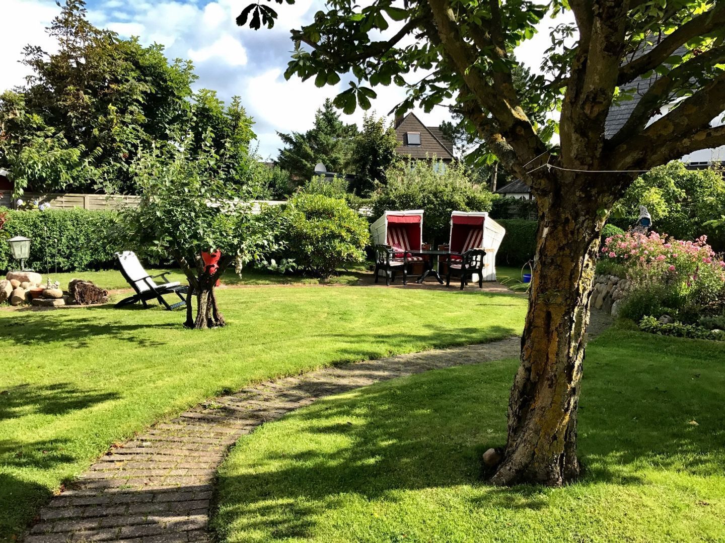 Ein gepflegter Garten mit einem gepflasterten Weg, einer Rasenfläche und zwei Sitzbereichen im Freien mit roten Markisen.