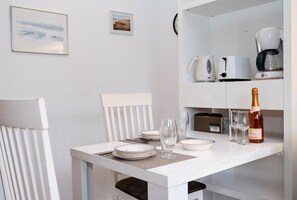 Une table à manger blanche, avec des assiettes, des verres et une bouteille de champagne. Deux tableaux encadrés sont accrochés au mur, ainsi qu’une chaise blanche et un meuble blanc contenant une machine à café et une radio.