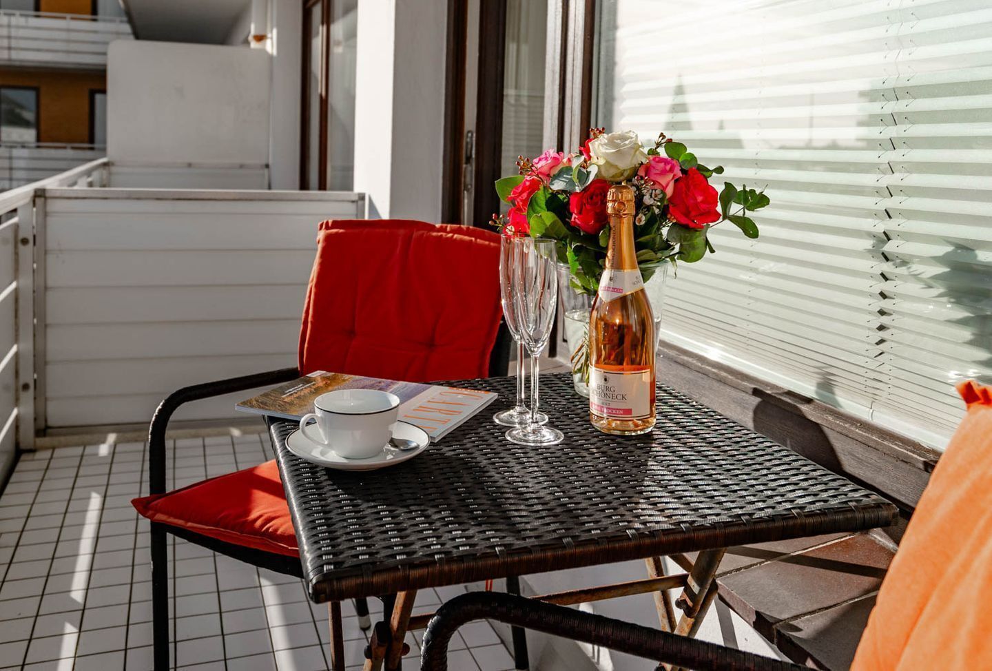 Et spisebord med vinflaske, et glas og en menu på en balkon med en rød stol og et hvidt borddække.