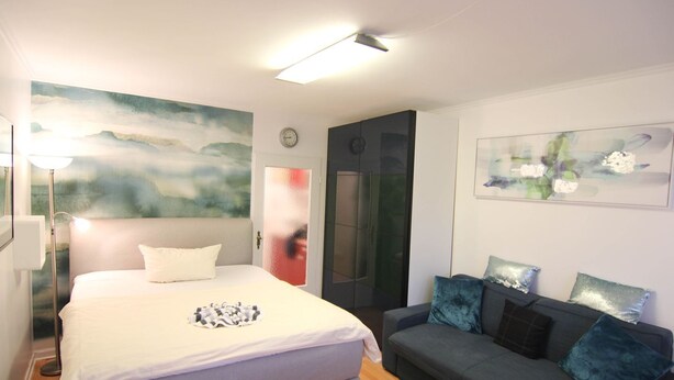 A bedroom with a bed, a grey sofa with cushions, a painting on the wall, and a door with a frosted glass panel. - Koje16 - Very cozy, adorably small and yet perfectly equipped. (Sylt)