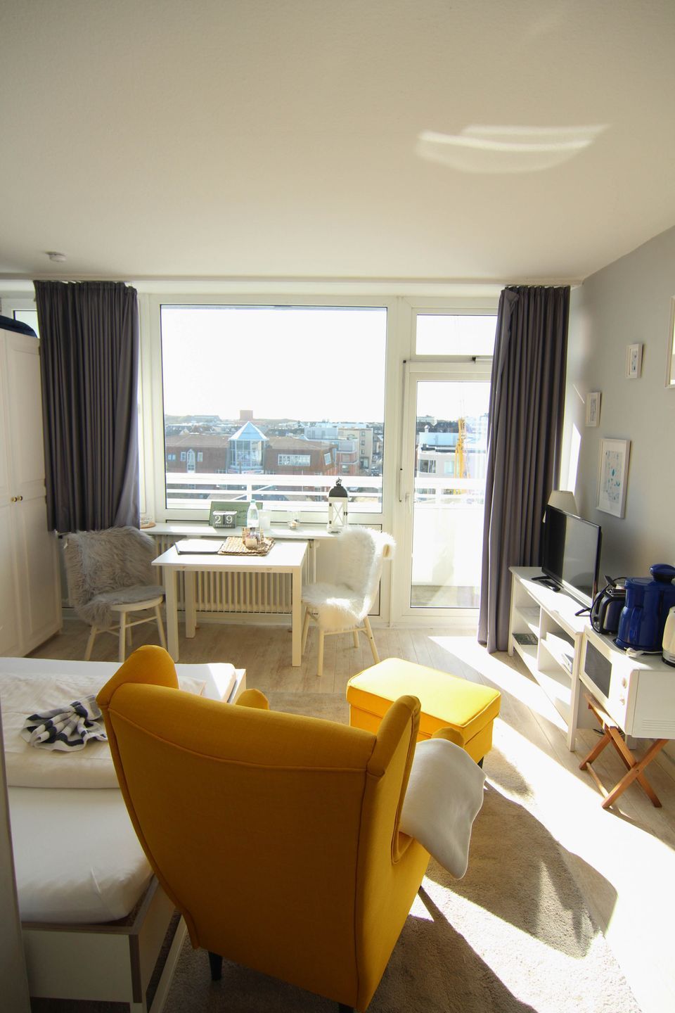Et hotelværelse med en gul lænestol, en hvid sofa, et lille bord, et fjernsyn og udsigt til bygninger gennem store vinduer.