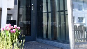 Entrada de un edificio moderno con puerta de vidrio, un letrero con el número '19' y un pequeño macizo de flores rosas.