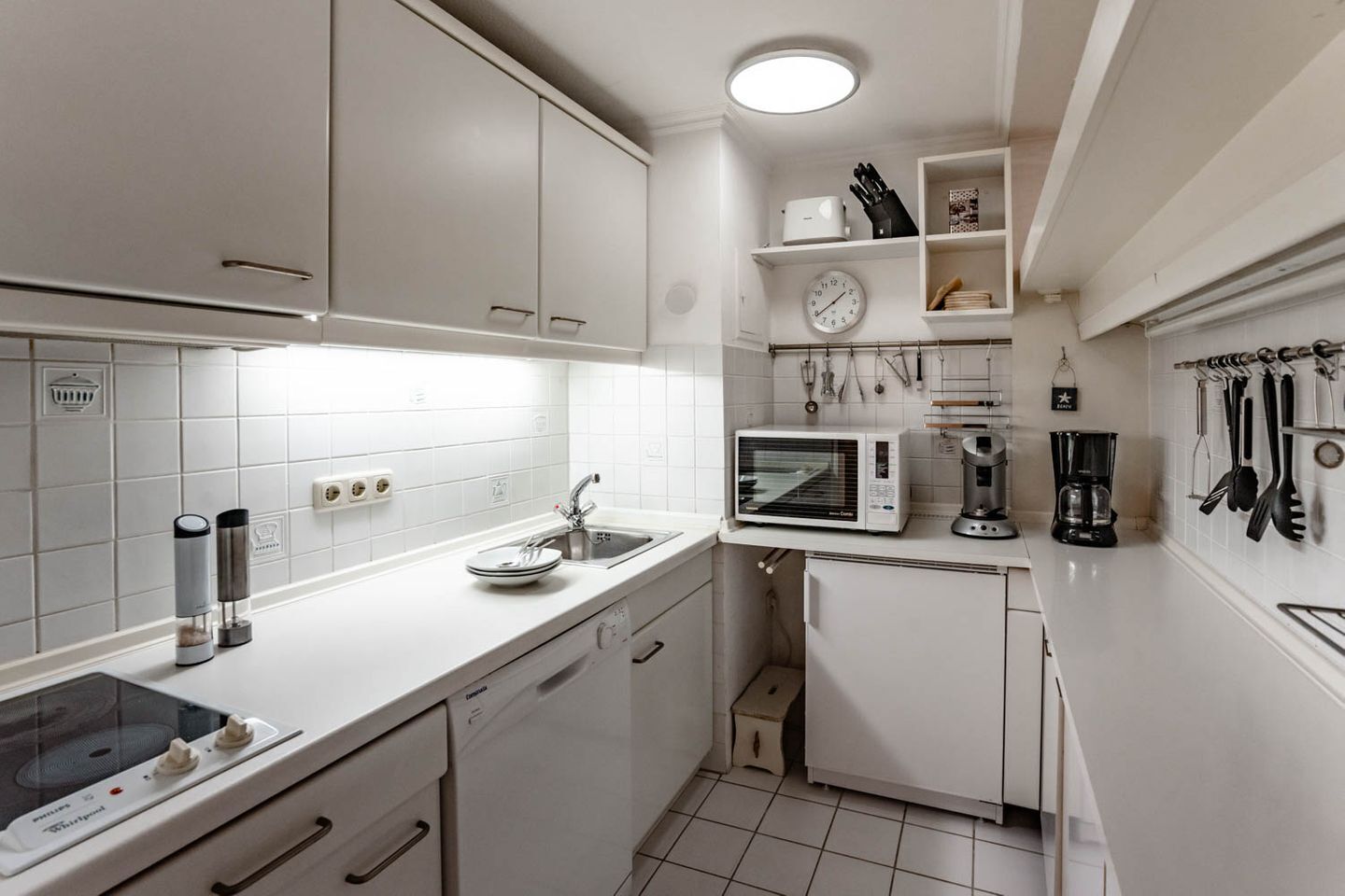 Una cocina compacta con armarios blancos, un microondas, una cafetera y diversos utensilios colgados en la pared.
