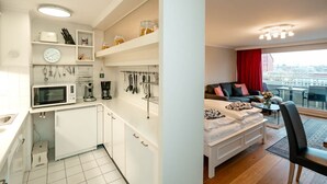 Une cuisine moderne avec des meubles blancs, un four à micro-ondes et divers ustensiles. À côté se trouve un salon avec un canapé, une table basse et une vue sur la ville.