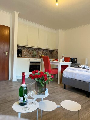 A hotel room with a bed, a dining table with chairs, a kitchen area, and a small white table with a bottle of champagne and glasses.